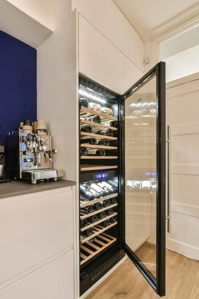 Buzdolabının arkasındaki ahşap döşemeli ve mavi duvarlı bir mutfakta şarap soğutucusu açık bir kapıdır.