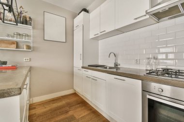 Sobanın üstündeki duvarda beyaz dolapları ve aletleri olan bir mutfak, köşede lavabo ve bulaşık makinesi.