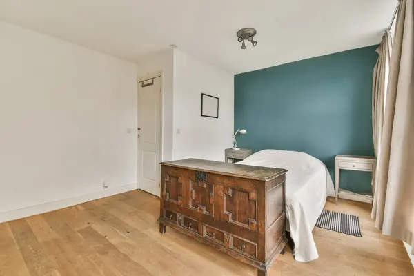 an empty bedroom with teal blue walls and wooden flooring the room has a bed, dresser, mirror and lamp