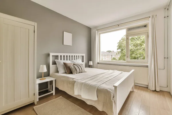 a bedroom with grey walls and hardwood flooring, white bed in the room has a large window overlooking out onto the street