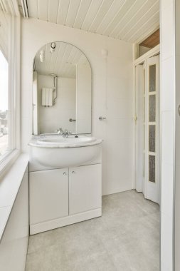 Köşesinde lavabo ve ayna olan beyaz bir banyo. Pencereye açılan açık bir kapının yanında.