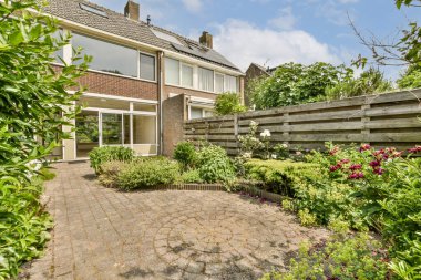 Ön planda bitki ve çiçeklerin olduğu bir dış alan. Arkasından alınan tuğladan bir kaldırım yolu.