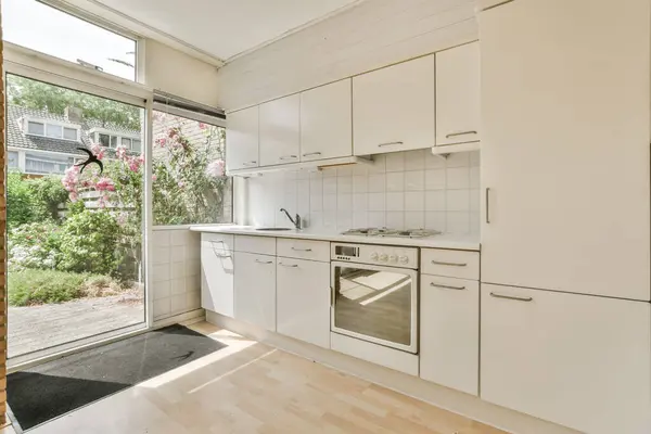 a kitchen area with white cupboards and appliances in the middle part of the room looking out to the garden