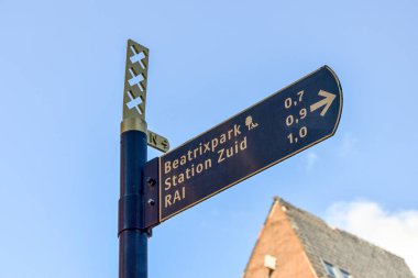 Mavi gökyüzü ve beyaz bulutlu tuğla bir binanın önündeki sokak tabelasının arkasında soldan sağa bakan bir ok var.