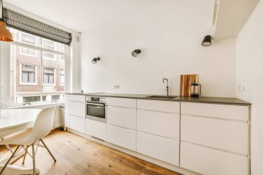 Beyaz dolapları, ahşap döşemeleri ve sokağa bakan açık bir penceresi olan bir evde mutfak ve yemek odası.