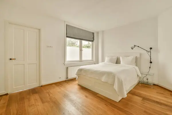 a bedroom with wood floors and white walls, including a large bed in the corner of the room on the right is a window