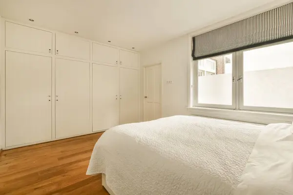 a bedroom with wood flooring and white cupboards on either side of the bed in front of the window