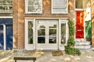 Beyaz kapılı bir tuğla bina ve binanın girişinin önündeki kaldırımda yeşil bitkiler büyüyor.