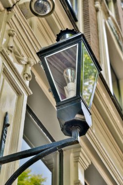 Arka planda ağaçlar ve pencereye yansıyan mavi gökyüzü olan bir binanın yanında asılı bir sokak lambası.