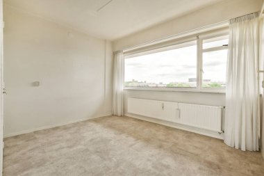 Beyaz duvarları ve yerde bej halısı olan boş bir oda. Şehre bakan bir pencere var.