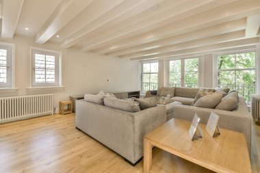 Ahşap döşemesi ve tavanında beyaz süslemeleri olan bir oturma odası. Dışarıdaki ağaçlara bakan pencereler.