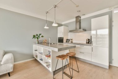 Beyaz dolapları, açık gri duvarları ve ahşap zemini olan bir dairede mutfak ve oturma odası çok temiz.