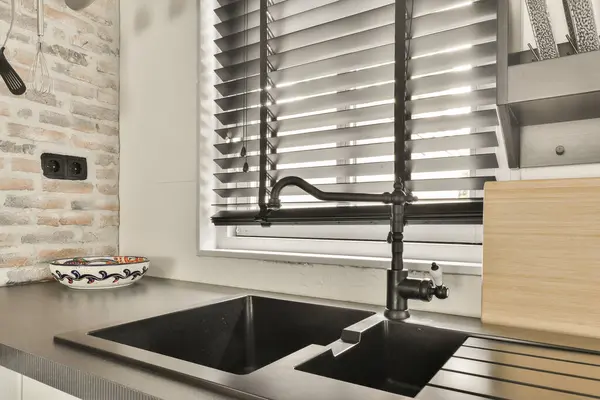 a kitchen sink in front of a window with shutters on the side and a bowl of fruit next to it