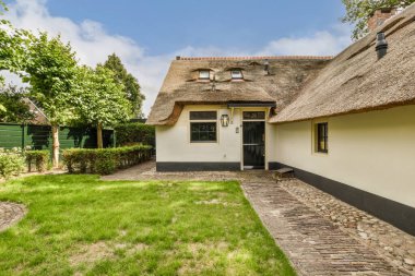 Mavi gökyüzünde yemyeşil ağaçlarla çevrili sazdan çatılı ve taş yolu ön kapıya uzanan bir ev.