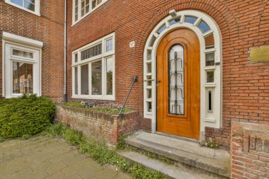 Ön bahçesinde yeşil bitkilerle çevrili beyaz çerçeveli ve pencereli kırmızı tuğlalı bir bina.