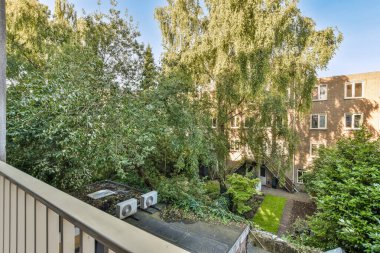 Ön tarafında ağaçlar ve çalılar olan bir apartman dairesi. Görüntü dışarıdan alınıp sokağa bakılıyor.