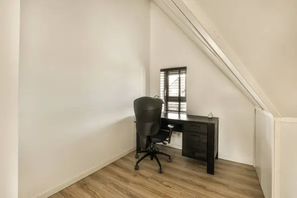 a desk and chair in the corner of a room with white walls, wooden floors and an attic style window