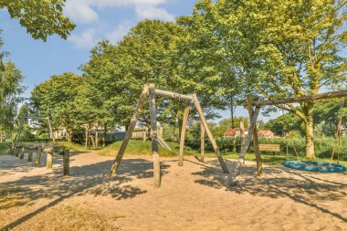 Ağaçlarla çevrili bir açık hava oyun alanında salıncaklı, slaytlı ve kumda kayan bir oyun alanı.