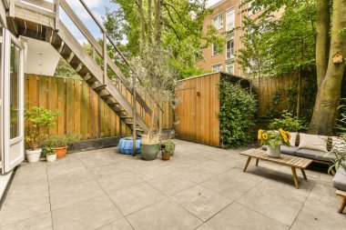 Evin yan tarafında ağaçlar ve bitkiler olan bir arka bahçe alanı. Burası açık hava yaşam alanı olarak kullanılıyor.