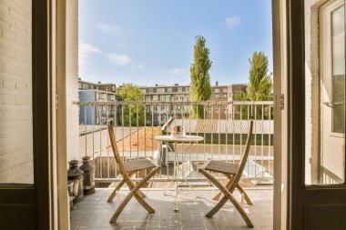 İki sandalyeli bir balkon ve ortada bir masa. Açık bir kapının içinden bahçeye bakıyor.