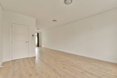 Beyaz duvarları ve sağ tarafında ahşap döşemeleri olan boş bir oda. Başka bir odaya açılan bir kapı var.