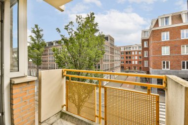 Arka planda ağaçlar ve binalar olan bir balkon. Bir apartmanın penceresinden sokağa bakıyor.