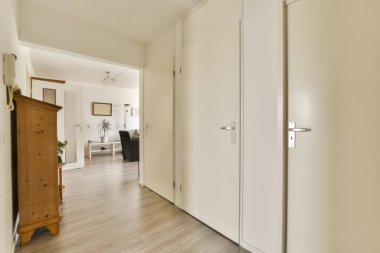 Odanın ortasında beyaz duvarları ve ahşap döşemeleri olan boş bir oda, başka bir odaya açılan bir kapı var.