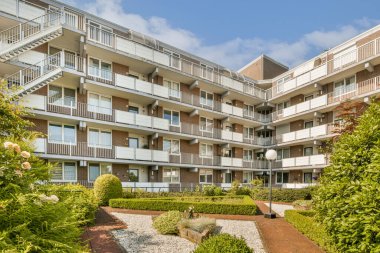 Parlak güneşli bir günde ön planda bitkiler ve çalılarla dolu bir apartman kompleksi, dışarıdan görüldüğü gibi.