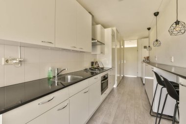 Fotoğrafın ortasında beyaz dolapları ve siyah tezgahları olan bir mutfak kapı girişinden çekiliyor.