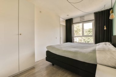 Penceresinde siyah perdeler ve beyaz yataklı bir yatak odası uzaktaki ağaçlara bakıyor.