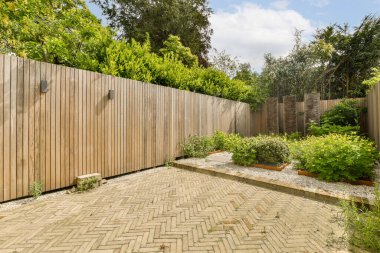 Arka bahçeye uzanan ahşap çitli ve tuğladan yollu bir bahçe. Mavi gökyüzü arka planında yemyeşil ağaçlarla çevrili.