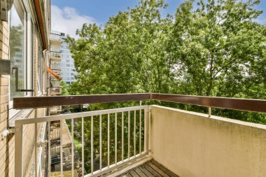 Arka planda ağaçlarla dolu bir balkon ve yukarıdaki mavi gökyüzü, güneşli bir günde bir apartmanın penceresinden dışarı bakarken görüldüğü gibi.