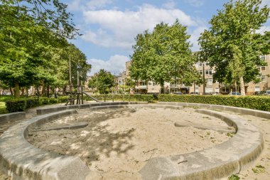 Güneşli bir günde, arka planda ağaçların ve binaların olduğu bir parkın ortasında bir kum çukuru.