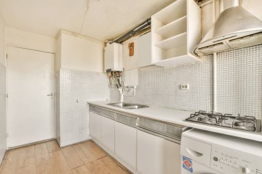 Çamaşır makinesi, bulaşık makinesi ve kurutma makinesinin önündeki tezgahın üstünde beyaz dolaplar ve aletler olan küçük bir mutfak.
