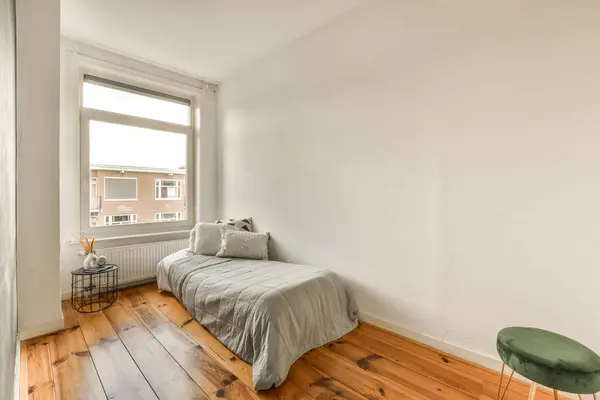 a bedroom with wood flooring and a bed in the corner, next to a window that looks out onto the street