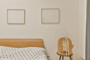 Beyaz bir yatak odasındaki yatağın üstündeki duvarda asılı iki çerçeveli resim. Siyah ve gri desenli yatak örtüsü.