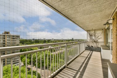 Arka planda ağaçlar ve binalar olan bir balkon, güneşli bir günde cam bir pencereden görüldüğü gibi.