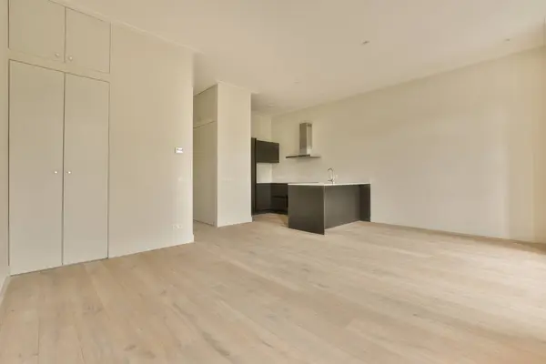 an empty living room with wood flooring and white cupboards on either side of the room there is a kitchen in the background