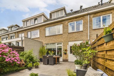 Ağaçlar ve çalılarla çevrili, tuğlalı bir binanın yanında çiçekler ve bitkiler olan bir dış alan.