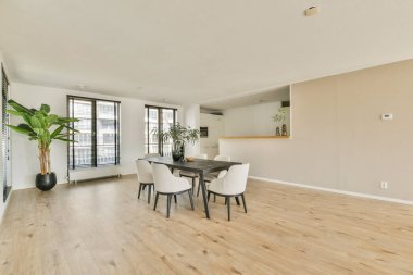 Ahşap bir masa, zarif sandalyeler ve pencerelerden süzülen doğal ışıkla aydınlatılmış büyük bir saksı bitkisinin yer aldığı modern bir yemek salonu..