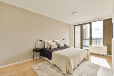 Tarafsız renk paleti, rahat yatak örtüsü ve doğal ışık sağlayan geniş pencereli modern bir yatak odası, huzurlu bir atmosfer yaratıyor..