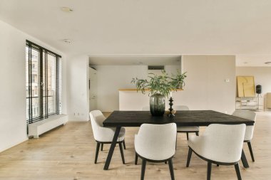 Gösterişli ve çağdaş bir yemek odası. Şık bir masa, sandalyeler ve boşluğu doğal ışıkla dolduran büyük pencereler..