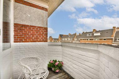 Beyaz duvarları olan büyüleyici bir balkon ve parlak çiçeklerle süslenmiş, berrak mavi gökyüzünün altındaki şık binaları gösteren bir sandalye..