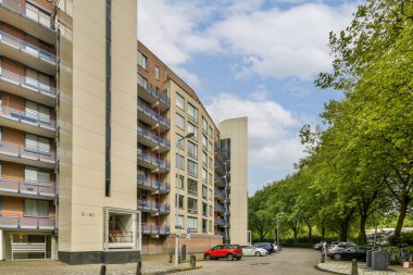 Çağdaş bir apartman kompleksi, iyi tasarlanmış mimarisi, yemyeşil ağaçlar ve açık mavi gökyüzü ile çevrili. Sahne, doğayla uyum içinde şehir yaşamını yakalıyor..