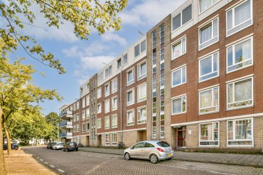 Geniş pencereleri ve hareketli dış görünüşü olan, ağaçlarla kaplı sakin bir caddenin yanında park halindeki arabaların bulunduğu çağdaş bir konut binası..