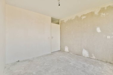 Beton bir duvar ve kapalı bir kapı içeren boş bir oda, minimalist tasarım unsurları ve tamamlanmamış yüzeyleri sergiliyor..