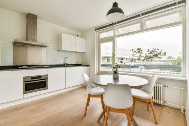 Zarif bir yemek masası, beyaz bir dolap ve doğal ışık alan geniş pencereli şık ve modern bir mutfak, davetkar bir yer yaratıyor..