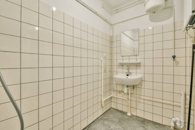 Minimalist bir banyo, fayanslı duvarlar, küçük bir lavabo ve bir ayna. Tasarım nötr renklerle işlevselliği ve temizliği vurgular.
