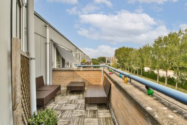 Hasır mobilyaların, güzel düzenlenmiş yeşilliklerin ve parlak gökyüzünün altındaki ağaç ve binaların manzarasının bulunduğu şık bir balkon..