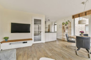 Duvara monte edilmiş TV, hafif ahşap döşeme ve canlı bitki dekoru olan şık ve modern bir oturma odası, rahat bir ortam yaratıyor..